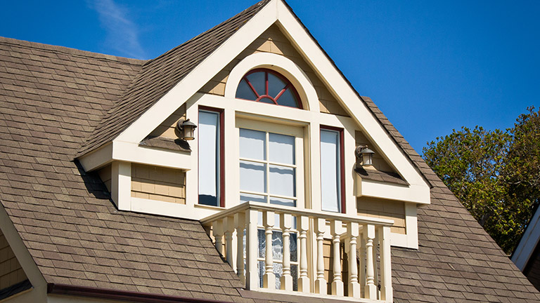 Gable dormer with a curved window and a small balcony extending from a brown shingle roof, illustrating a roof structure that could be susceptible to sagging if underlying issues exist.