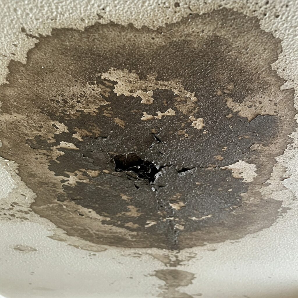 nterior view of a white ceiling with a large, circular, dark brown water stain and a hole in the center, indicating significant water leakage likely resulting from improper roof installation.